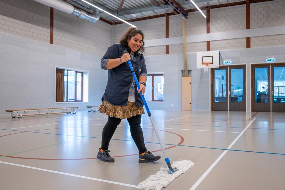 Een medewerker veegt met een grote bezem de sporthal aan